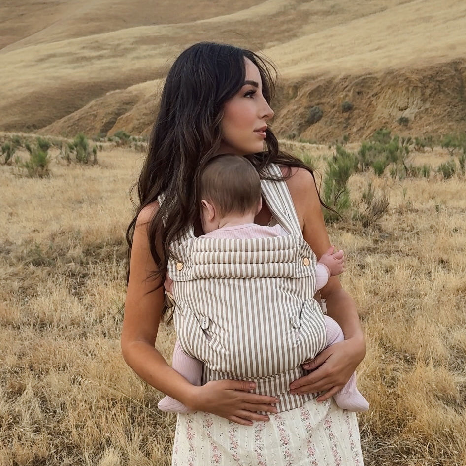 Woman carrying a baby in a striped baby carrier against a scenic background of hills and blue sky.